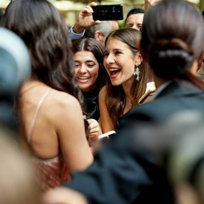 Event photographer point of view past cameras and with focus on enthusiastic admirers smiling and talking as they greet brunette woman in evening gown.